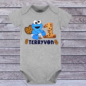 Pu&ograve; includere: Un body grigio per neonati con un personaggio dei cartoni animati blu, un biscotto e il numero 1. Il testo "TERRYVON" &egrave; stampato in blu navy. Il body &egrave; realizzato in materiale morbido e ha maniche corte.