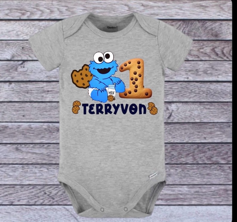 Pu&ograve; includere: Un body grigio per neonati con un personaggio dei cartoni animati blu, un biscotto e il numero 1. Il testo "TERRYVON" &egrave; stampato in blu navy. Il body &egrave; realizzato in materiale morbido e ha maniche corte.
