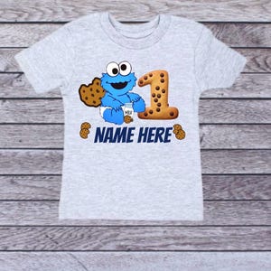Pu&ograve; includere: Una maglietta grigio chiaro con un personaggio dei cartoni animati blu che tiene in mano un cartone di latte e un biscotto. Il numero "1" &egrave; disegnato come un biscotto. Il testo "NAME HERE" &egrave; stampato in blu.