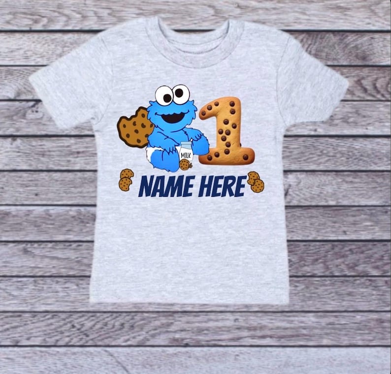 Pu&ograve; includere: Una maglietta grigio chiaro con un personaggio dei cartoni animati blu che tiene in mano un cartone di latte e un biscotto. Il numero "1" &egrave; disegnato come un biscotto. Il testo "NAME HERE" &egrave; stampato in blu.