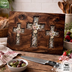 May include: A rectangular wooden board featuring three carved cross shapes filled with clear resin, embedded with dried flowers and small stones. The wood displays a dark, natural grain pattern with visible knots, and the floral arrangement includes white lilies and pink roses.