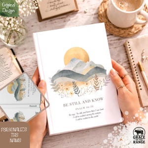 May include: A white journal with a watercolor design of mountains, a sun, and floral accents. The text "BE STILL AND KNOW" and Psalm 46:10 are printed on the cover. The journal is held open, revealing a personalized name.