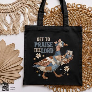 May include: A black tote bag featuring a graphic of a goose designed with various brown, blue, and white patchwork patterns. The goose holds a small book labeled Holy Bible. The text reads Off to Praise the Lord, surrounded by small flower and star icons.