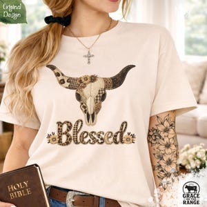 May include: Cream-colored t-shirt featuring a decorative bull skull design with patchwork patterns and floral accents. The word "Blessed" is written in a decorative font. The shirt also has the text "Original Design" and "Grace on the Range".