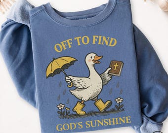 May include: A blue jean colored sweatshirt featuring a graphic of a white duck wearing yellow rain boots, holding a yellow umbrella and a brown book with a cross on the cover. The text reads OFF TO FIND GOD'S SUNSHINE in yellow lettering.
