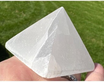 Puede incluir: Un cristal transparente en forma de pirámide con una superficie lisa y pulida. El cristal es de un blanco pálido y translúcido y se sostiene en una mano. El fondo es un césped verde borroso.