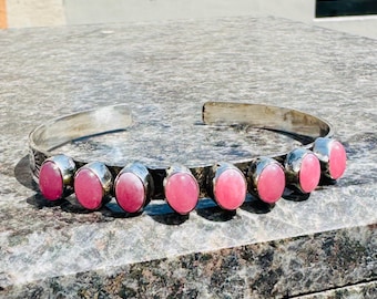 Puede incluir: Un brazalete de plata con siete piedras preciosas rosas ovaladas. El brazalete tiene un diseño abierto simple, con las piedras engastadas en biseles de plata. El brazalete se exhibe sobre una superficie gris texturizada.