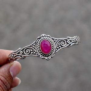 May include: A silver-toned cuff bracelet featuring an oval-shaped, vibrant pink cabochon stone at the center. The metal band is decorated with intricate, raised scrollwork and rope-like detailing surrounding the central pink gemstone.