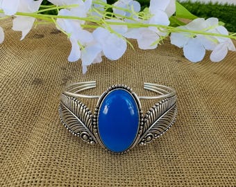 May include: A silver-toned cuff bracelet featuring a large, smooth, oval-shaped blue stone at the centre. The metal band is decorated with detailed, embossed feather patterns on both sides of the central stone, creating a symmetrical design.