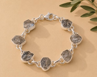 May include: A silver chain bracelet featuring six irregular, faceted gray stones set in polished silver bezels. The bracelet is constructed with circular silver links connecting each stone setting and includes a silver lobster claw clasp closure.