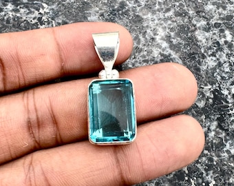 May include: A rectangular, light blue gemstone pendant with a silver bail. The gemstone is set in a silver bezel and has a clear, transparent appearance. The pendant is held in a hand against a textured, dark background.
