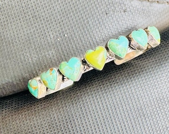 Puede incluir: Horquilla plateada con piedras turquesas y amarillas en forma de corazón. La horquilla tiene siete piedras en forma de corazón, con una mezcla de colores turquesa y amarillo. La horquilla está sobre un fondo texturizado.