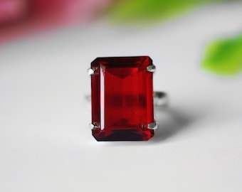 Könnte beinhalten: Ein silberner Ring mit einem großen, rechteckigen, tiefroten Edelstein. Der Edelstein ist der Mittelpunkt, gefasst in einem schlichten, eleganten Silberband. Der Ring wird vor einem weichen, verschwommenen Hintergrund gezeigt.