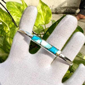 Könnte beinhalten: Ein silbernes Armband mit einem rechteckigen Inlay aus türkis- und weißfarbenem Stein. Das Armband hat eine polierte Oberfläche. Im Hintergrund sind grüne Blätter zu sehen.