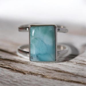 May include: A silver ring with a rectangular, light blue gemstone. The ring has a double-band design, with the gemstone set in a silver bezel. The gemstone has a marbled appearance, with shades of blue and white.