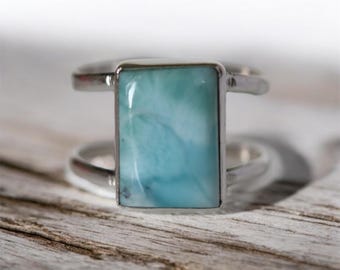 May include: A silver ring with a rectangular, light blue gemstone. The ring has a double-band design, with the gemstone set in a silver bezel. The gemstone has a marbled appearance, with shades of blue and white.