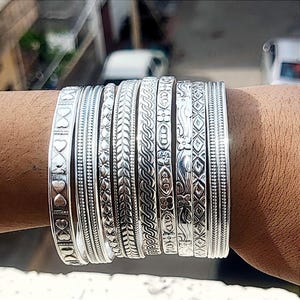 May include: A collection of silver bangles with various embossed designs, including hearts, leaves, and geometric patterns. The bangles are stacked on a wrist, showcasing their intricate details and reflective surface.