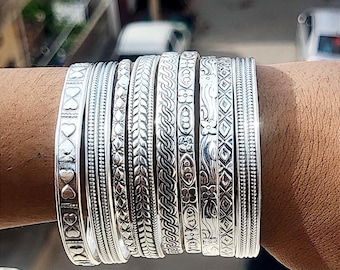 Puede incluir: Una colección de pulseras de plata con varios diseños en relieve, incluyendo corazones, hojas y patrones geométricos. Las pulseras están apiladas en una muñeca, mostrando sus intrincados detalles y superficie reflectante.