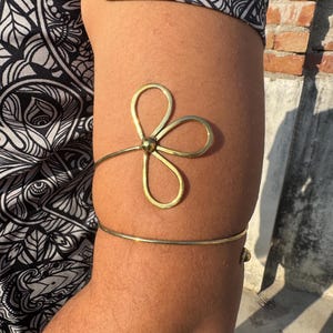 May include: A gold-colored arm cuff featuring a floral design. The cuff has a simple band that wraps around the upper arm, with a delicate flower accent. The flower has three petals and a central sphere.