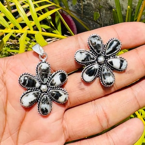 May include: A flower-shaped pendant and matching stud earrings. The jewelry features a silver-toned metal setting with black and white stone petals. The pendant has a loop for a chain. The earrings and pendant are displayed on a hand.