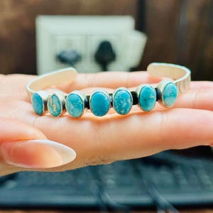 May include: Silver cuff bracelet featuring seven oval turquoise stones. The stones are set in individual bezels and are a vibrant blue-green color. The bracelet has a simple, elegant design, perfect for everyday wear.