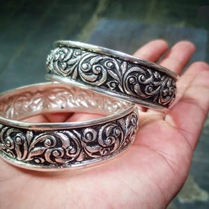Puede incluir: Dos pulseras de plata con intrincados adornos florales. Las pulseras son anchas, con un patrón detallado de hojas y espirales. El metal tiene un aspecto ligeramente oxidado, realzando la profundidad y textura del diseño. Las pulseras se muestran en una mano.
