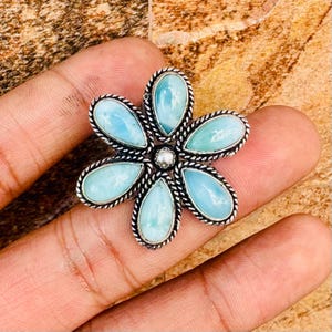 May include: A silver-toned ring featuring a flower design with six light blue, teardrop-shaped stones. The ring has a beaded border and a small, round silver center stone. The ring is set against a neutral background.