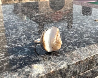 May include: A silver ring featuring a large, irregularly shaped, natural stone. The stone has a light beige and white color palette with a polished surface. The ring is set on a dark, reflective surface.