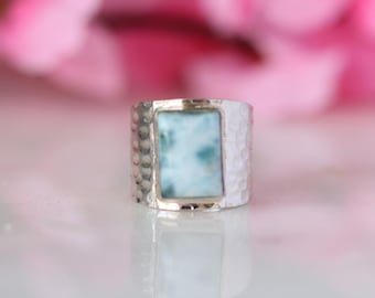May include: A silver ring featuring a rectangular, pale blue gemstone. The ring's band has a hammered texture. The gemstone displays a marbled pattern with white and blue tones. The background is blurred with pink and white hues.