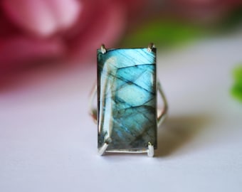May include: A rectangular labradorite gemstone ring. The gemstone has a blue and green iridescent shimmer. The ring's band is silver-colored. The background is blurred, with pink and green hues.
