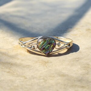 May include: A silver-toned bracelet with a heart-shaped abalone shell center. The iridescent shell features shades of green, blue, and purple. The bracelet has a delicate, openwork design with curved lines and small decorative spheres.