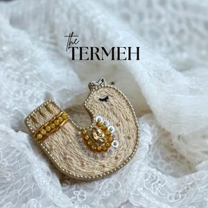 Peut inclure: Une broche beige faite à la main avec des perles et des broderies complexes. La broche présente un motif en forme d'oiseau avec des accents dorés et blancs, et le texte "the TERMEH" est visible en arrière-plan. La broche est posée sur un tissu en dentelle blanche.