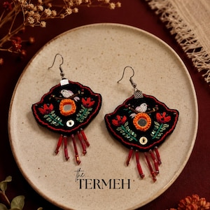 May include: Pair of black embroidered earrings with red and orange floral designs, a small mirror, and dangling red beads. The earrings are fan-shaped and have silver-colored hooks. The earrings are displayed on a beige plate.