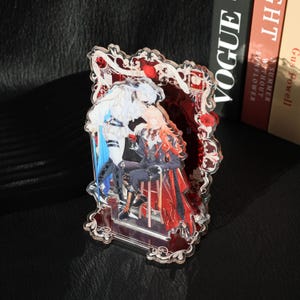 May include: A decorative acrylic stand featuring a romantic illustration of two figures in an ornate frame. The figures are depicted in shades of white, blue, and red, with intricate details and a vintage aesthetic. The stand is clear with a decorative border.