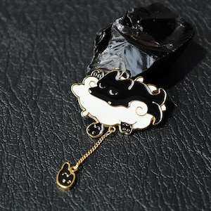 May include: A black and white enamel pin in the shape of a cat on a cloud, with a gold chain and a small cat charm. The pin has a gold border and is set against a dark background.