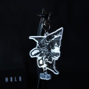 May include: A clear acrylic keychain featuring a black and white illustration of a character with shark-like features, including a shark head hood. A small rectangular tag attached at the bottom displays the text Ellen Joe, with a star-shaped metal clasp.