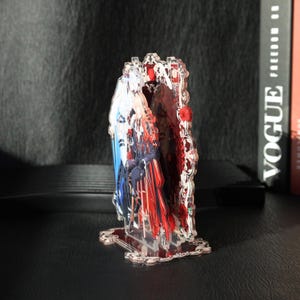 May include: A clear acrylic decorative piece with a detailed design. The piece features a central image of a person in red and blue attire, framed by a decorative border with red accents. The base and border have a floral design.