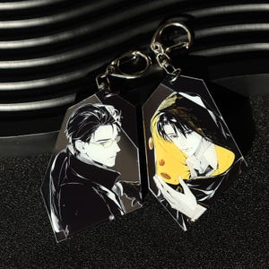 May include: Two anime-style keychains featuring male characters. Each keychain has a black and white illustration of a man. One man wears glasses and a coat. The other is hooded with a yellow interior. Both have silver key rings.