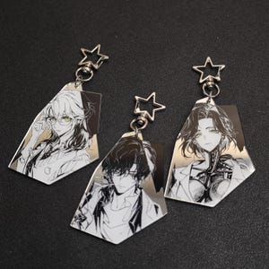 Puede incluir: Tres llaveros de personajes de estilo anime. Cada llavero presenta una ilustración en blanco y negro de una persona con un cierre en forma de estrella. Los llaveros tienen un acabado metálico plateado y se muestran sobre una superficie oscura.