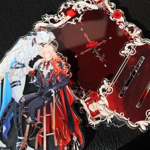 May include: Acrylic charms featuring anime-style illustrations. One charm depicts two figures, one in white and blue, the other in a black suit with red accents. The other charm is a dark red design with roses and decorative elements.