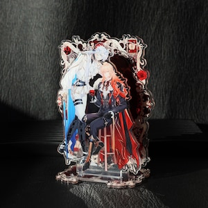 以下が含まれることがあります： 2人のアニメ風のキャラクターが描かれた透明なアクリルスタンド。一人は青いアクセントの白い衣装、もう一人は赤いマントの黒いスーツを着ています。スタンドにはバラの装飾フレームが付いています。