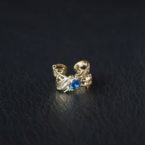 Puede incluir: Un brazalete de oreja dorado con un diseño abierto que presenta intrincados detalles de hojas y vides. Una vibrante piedra preciosa azul está engastada en el centro del brazalete. La joya se muestra sobre un fondo oscuro.