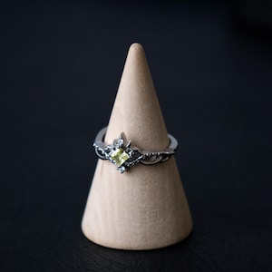 Peut inclure: Bague en argent ornée d'une pierre précieuse carrée jaune clair. La bague présente un motif décoratif autour de la pierre et le long de l'anneau. Elle est présentée sur un présentoir conique marron, sur fond sombre.
