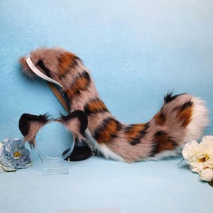 May include: A costume set with a spotted tail and ear headband. The tail is brown, orange, and black with a white tip. The ears are black with brown fur inside. The set is made of faux fur and is suitable for cosplay or costume events.