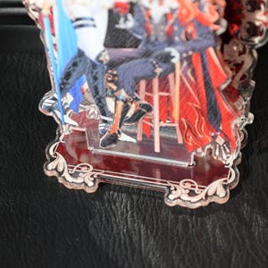 May include: A clear acrylic stand featuring a colorful illustration of two figures. The stand has a decorative base with a red border and ornate scrollwork. The figures are dressed in detailed costumes with red, blue, and black accents.