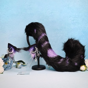 Può includere: Un set di coda e fascia per orecchie da gatto in finta pelliccia nera e viola. Le orecchie hanno accenti viola e ciondoli pendenti argentati con perle finte. La coda è nera con strisce viola.