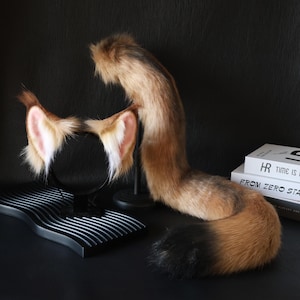 Puede incluir: Un conjunto de cola de zorro de piel sintética marrón y negra y diadema de orejas a juego. Las orejas son de color canela con detalles rosas en el interior. La cola es un degradado de marrón a negro. El conjunto está diseñado para cosplay o disfraces.