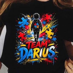 May include: Black t-shirt with a colorful graphic design. The design features a child walking forward, surrounded by puzzle pieces in red, blue, and yellow. The text "TEAM DARIUS" is prominently displayed in blue and yellow.