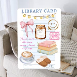 Puede incluir: Un diseño decorativo de tarjeta de biblioteca con un lazo rosa, una vela encendida, una pila de libros, una taza de café y zapatillas. La tarjeta incluye el texto "LIBRARY CARD" y la cita "True love stories never have endings."