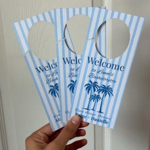 May include: Three vertical door hangers featuring a light blue and white striped pattern. Each hanger has a circular cutout at the top and displays the text Welcome to Lenni's Bachelorette, Tybee Island, Georgia, and the names Paige + Danielle.